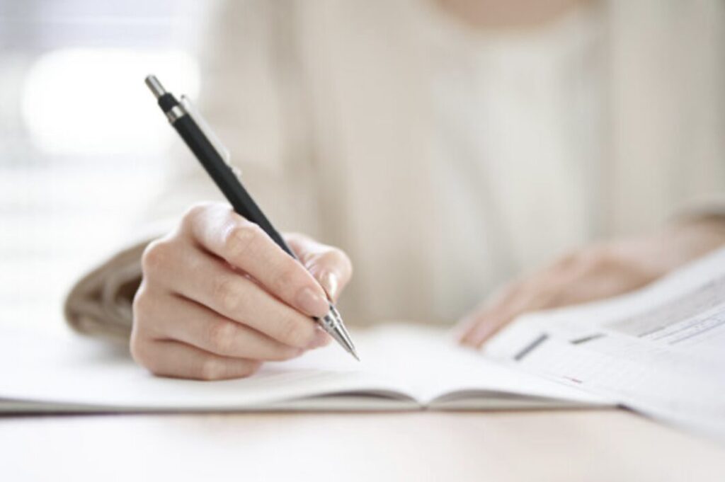 ノートにペンで書き込みながら勉強している様子。白い机の上で問題集を開き、手元に集中しているシーン。