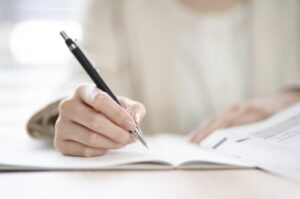 ノートにペンで書き込みながら勉強している様子。白い机の上で問題集を開き、手元に集中しているシーン。