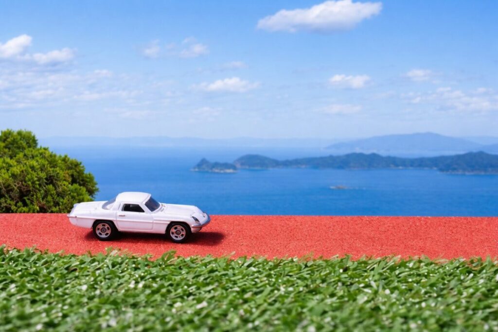 白いクラシックカーのミニチュア模型が赤い道の上を走り、背景に青い海と島々の景色が広がる風景。