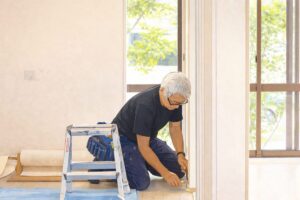 室内で脚立の横に膝をつき、窓際のドア枠に仕上げ作業を行う職人の様子を描いたイラスト